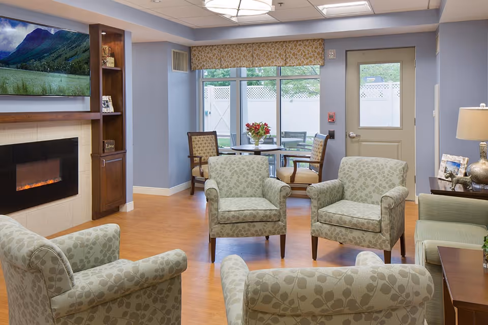 A cozy senior living common area with four patterned armchairs arranged around a wooden coffee table. A flat-screen TV is mounted above a modern electric fireplace on the left wall. In the background, there is a small round table with four chairs near large windows and a door leading outside. The room has light blue walls, wood flooring, and soft lighting from ceiling fixtures and a table lamp.