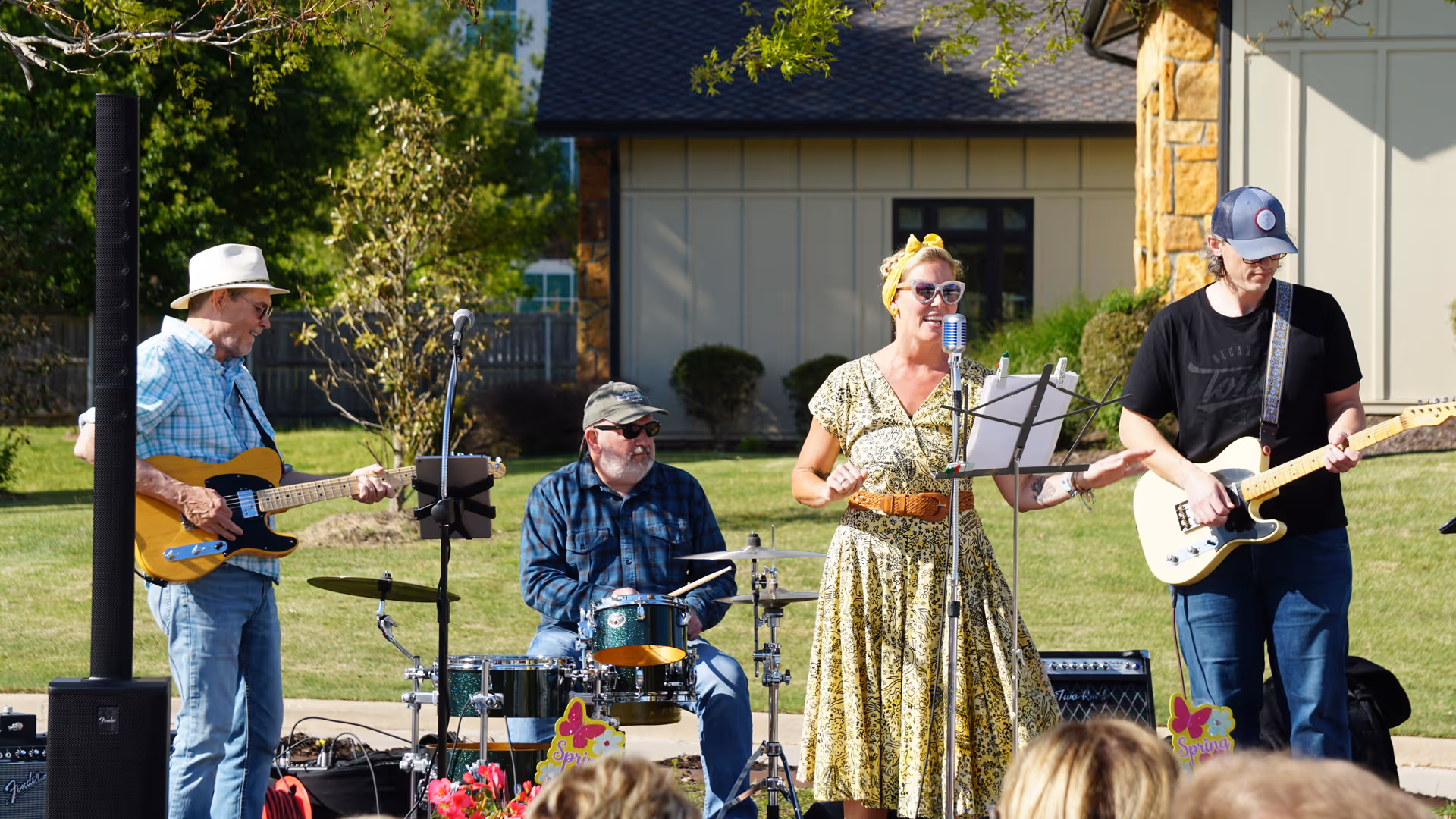 A small outdoor band performance with four musicians: two men playing electric guitars, one man playing drums, and a woman singing into a microphone. They are performing on a grassy area in front of a building with beige siding and stone accents. The woman is wearing a yellow patterned dress and sunglasses, and the men are casually dressed. There are some colorful spring-themed decorations near the drum set.