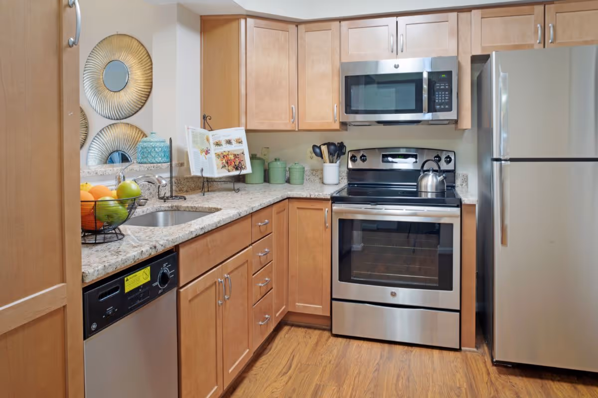 A modern kitchen featuring wooden cabinets, a granite countertop, a stainless steel sink, dishwasher, stove with oven, microwave, and refrigerator. Decorative items include a fruit basket, a cookbook on a stand, green canisters, and round metallic wall decor.