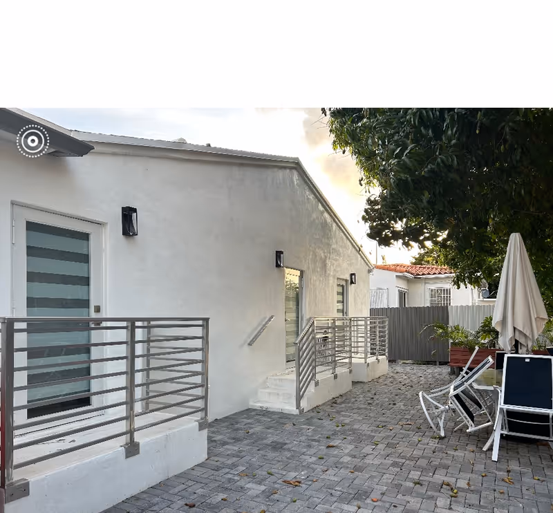 Outdoor patio area with a paved stone floor, white building walls with metal railings along steps and doors, several white lounge chairs, a closed white umbrella, and greenery including a large tree on the right side.