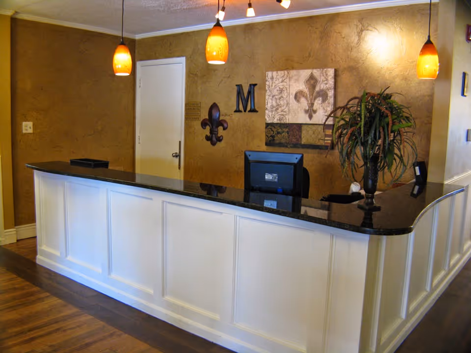 Reception desk area with a black countertop and white paneling. Three orange pendant lights hang from the ceiling above the desk. Behind the desk is a brown textured wall with decorative items including a large letter M, a fleur-de-lis wall decoration, and a framed artwork. A potted plant is placed on the right side of the desk.