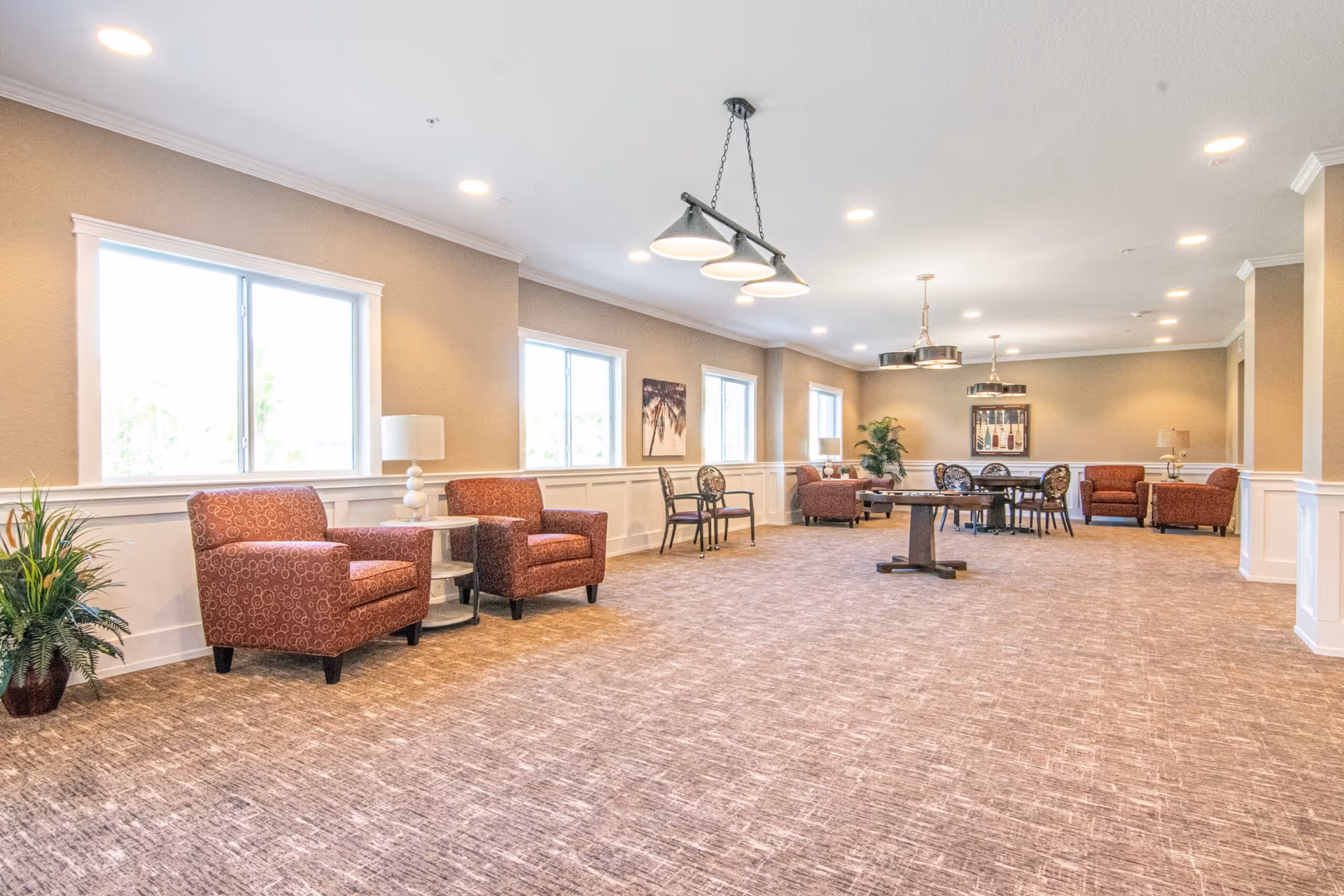 A spacious, well-lit common area with beige walls and carpeted floor. The room features several red patterned armchairs, small round tables with lamps, and a few black chairs with decorative backs. Large windows line one wall, allowing natural light to fill the space. There are framed artworks on the walls and potted plants adding greenery. Modern ceiling lights hang from the white ceiling.
