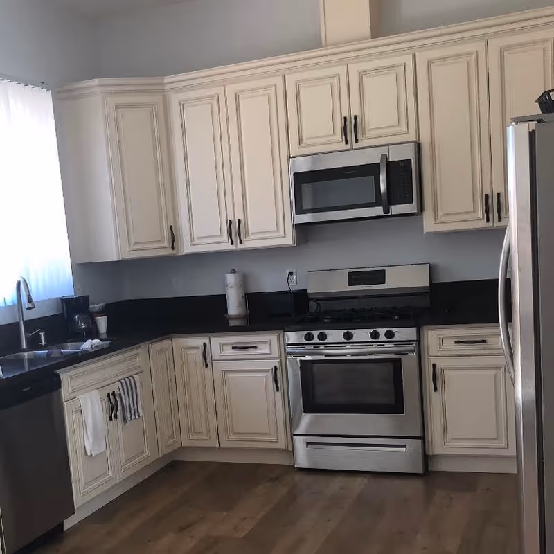 A modern kitchen with cream-colored cabinets, black countertops, and stainless steel appliances including a microwave, stove, dishwasher, and refrigerator. There is a window with sheer white curtains letting in natural light, a coffee maker on the counter, and two towels hanging on the cabinet handles.