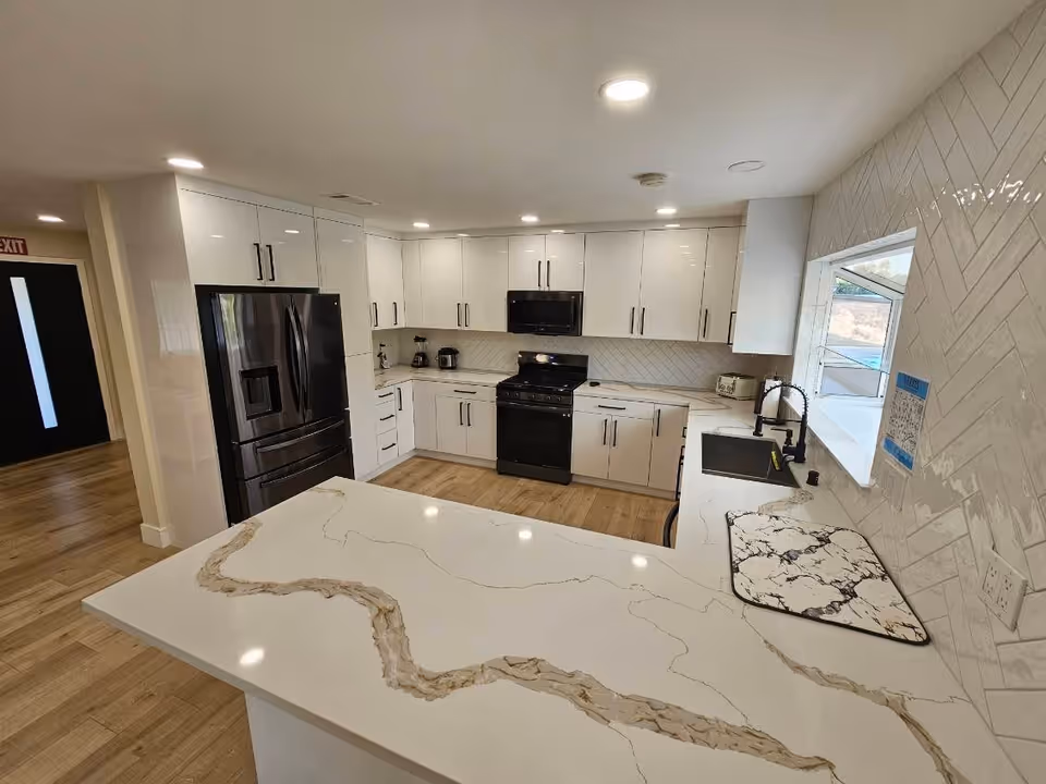Modern kitchen with white cabinets, black appliances including a refrigerator, stove, and microwave, a large marble countertop island, wood flooring, and a window above the sink.