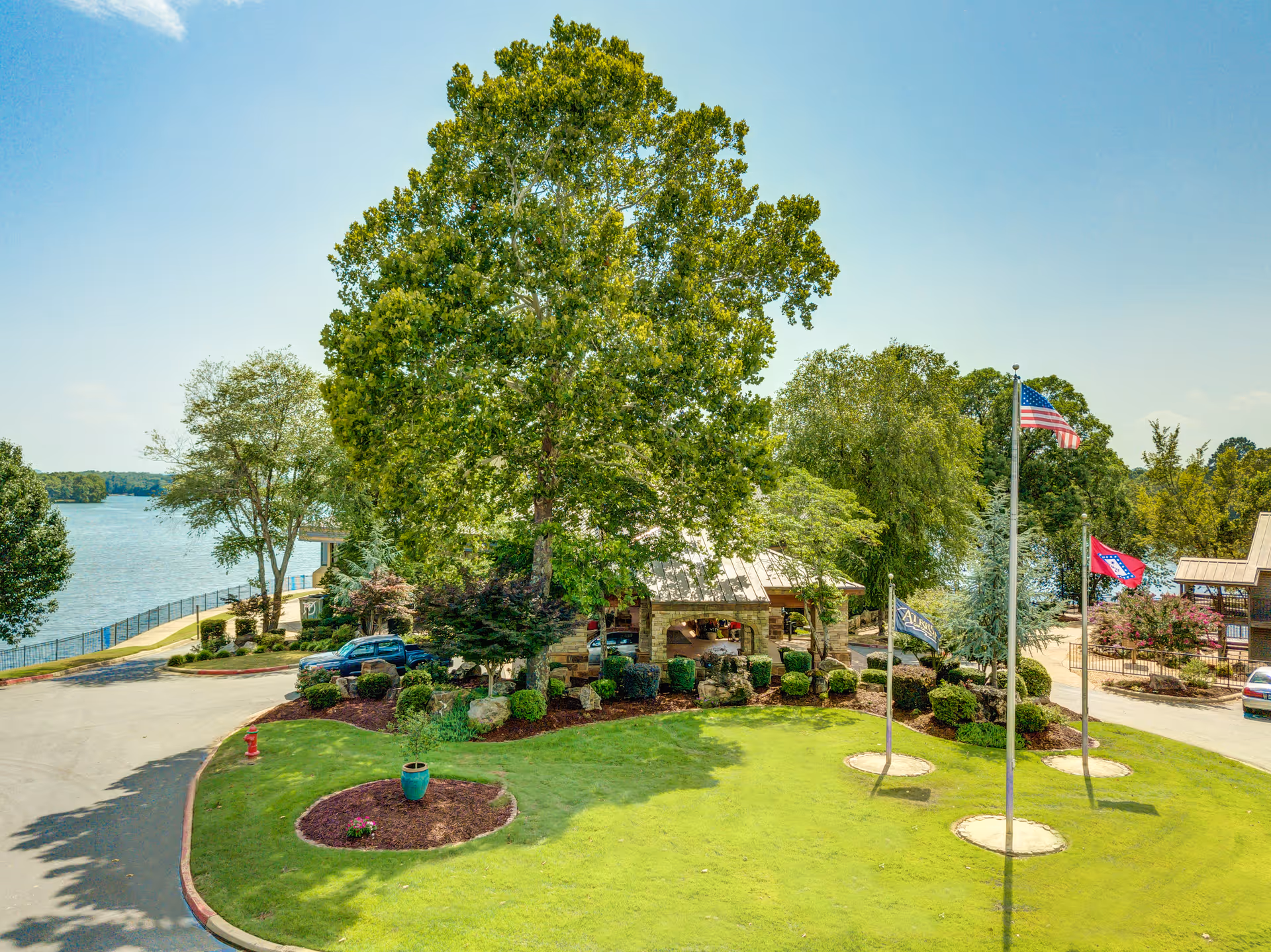 Landscaped entrance with a large tree, flagpoles, lawn and a covered pavilion beside a lake.
