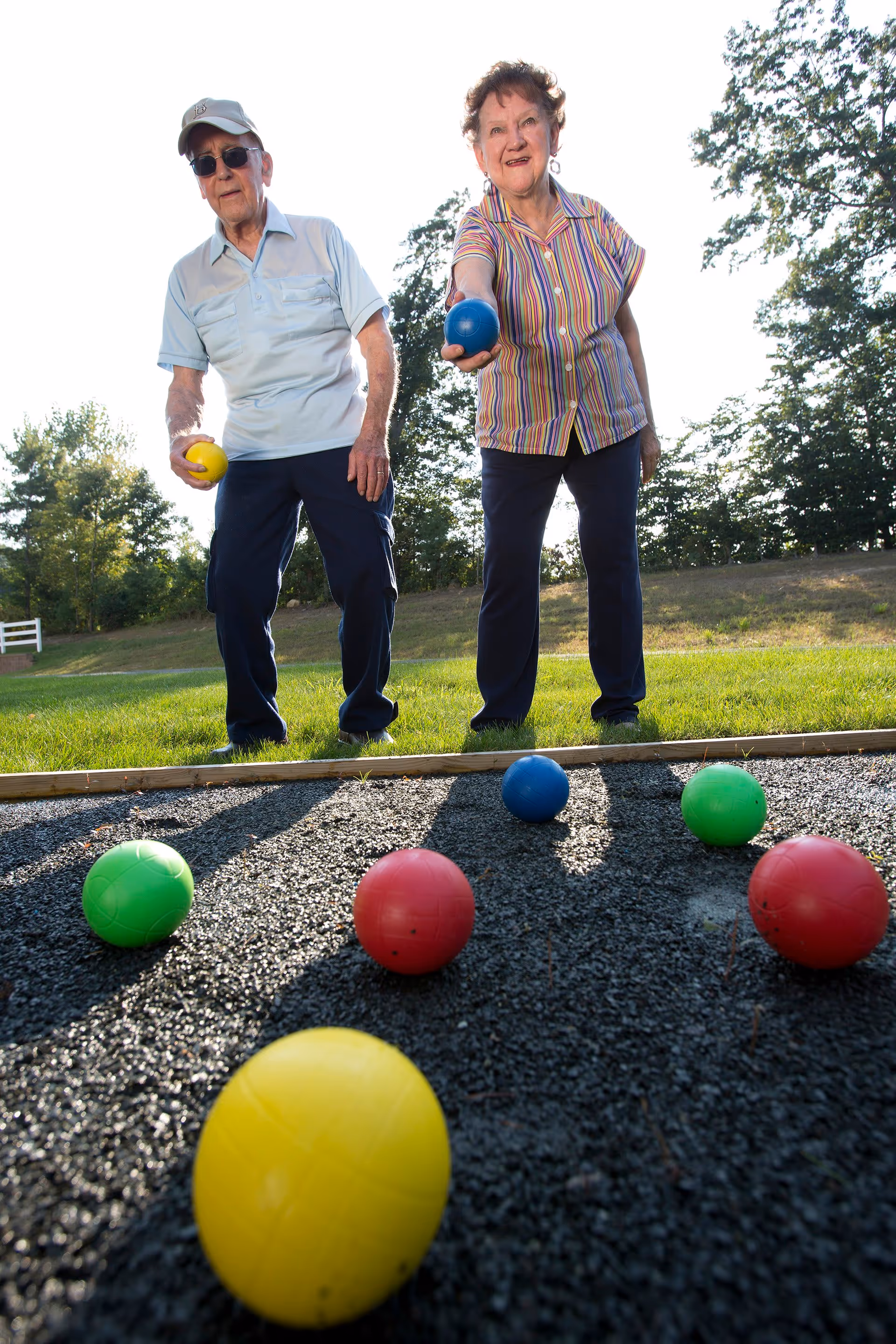 An elderly man and woman playing bocce ball outdoors on a sunny day. The man is wearing a light blue shirt, dark pants, a white cap, and sunglasses, holding a yellow ball. The woman is wearing a striped shirt and dark pants, holding a blue ball and smiling. Several colorful bocce balls are on the ground in front of them, with trees and grass in the background.