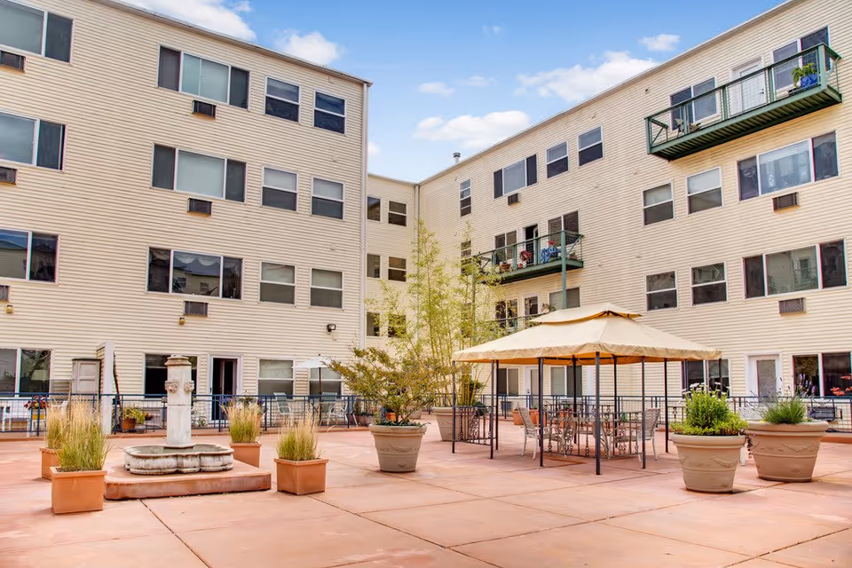 Courtyard with potted plants, a fountain and tables under a canopy surrounded by a multi-story beige apartment building.