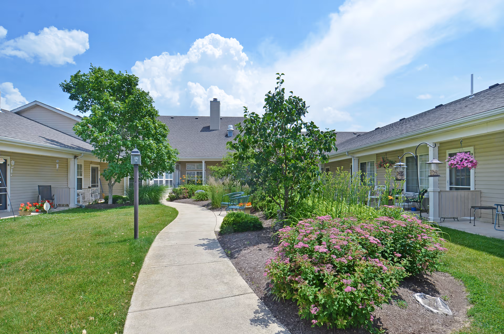 A sunny outdoor courtyard at Muncie Estates Senior Living featuring a concrete walkway winding through green grass, flowering bushes, and small trees. The courtyard is surrounded by single-story beige buildings with windows and patio chairs. The sky is blue with some clouds.