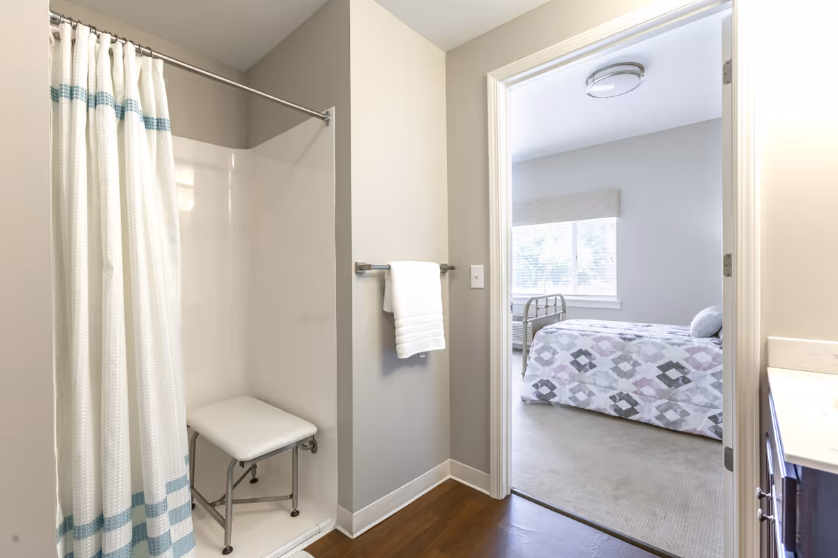 View of a bathroom with a shower area featuring a white curtain with blue stripes and a white shower chair inside. A white towel hangs on a towel rack on the wall. The bathroom opens into a bedroom with a bed covered in a patterned quilt and a window with blinds letting in natural light.