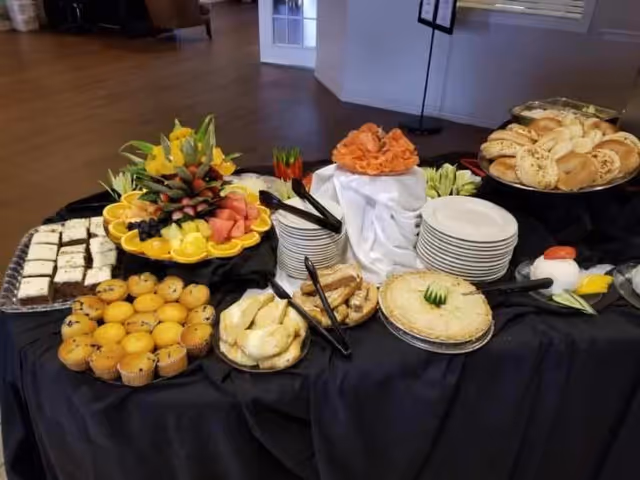 A buffet table set with a variety of foods including muffins, pastries, bagels, a fruit platter with watermelon, grapes, and pineapple, a plate of smoked salmon, and a pie. There are stacks of plates and serving utensils on a black tablecloth in a room with wooden flooring and a door in the background.