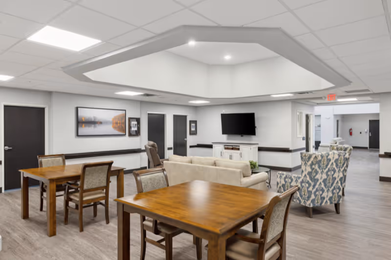 A bright communal lounge with wooden tables and chairs, a sofa and patterned armchairs facing a wall-mounted TV.