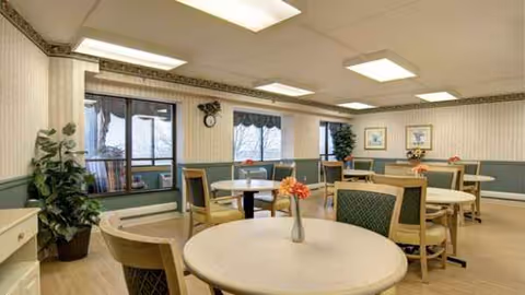 Bright dining room with round tables, chairs, plants, and framed art in a senior facility.