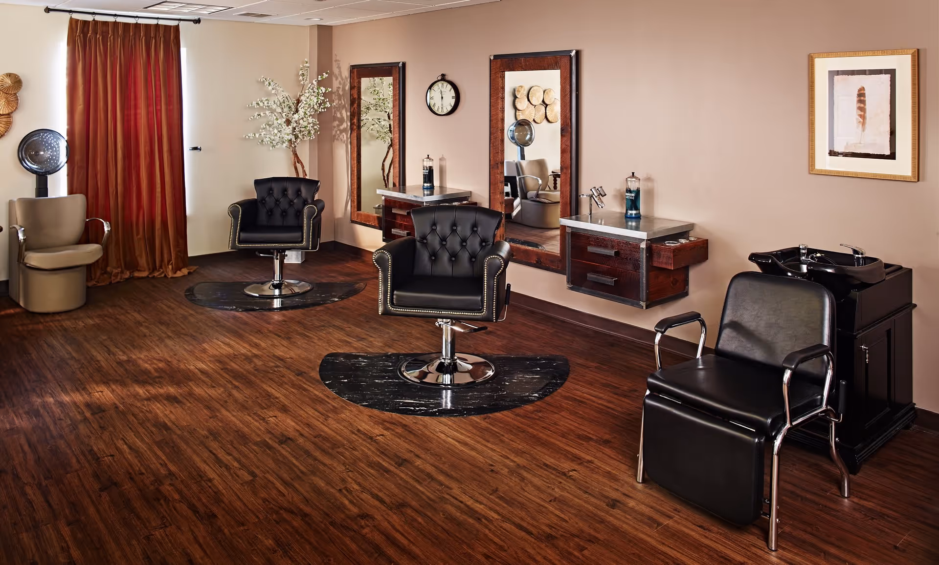 Interior of a salon area with two black salon chairs in front of large mirrors mounted on a beige wall. There is a black shampoo station chair with a sink on the right side and a beige chair with a hair dryer on the left side near red curtains. The floor is wooden and the room is softly lit.