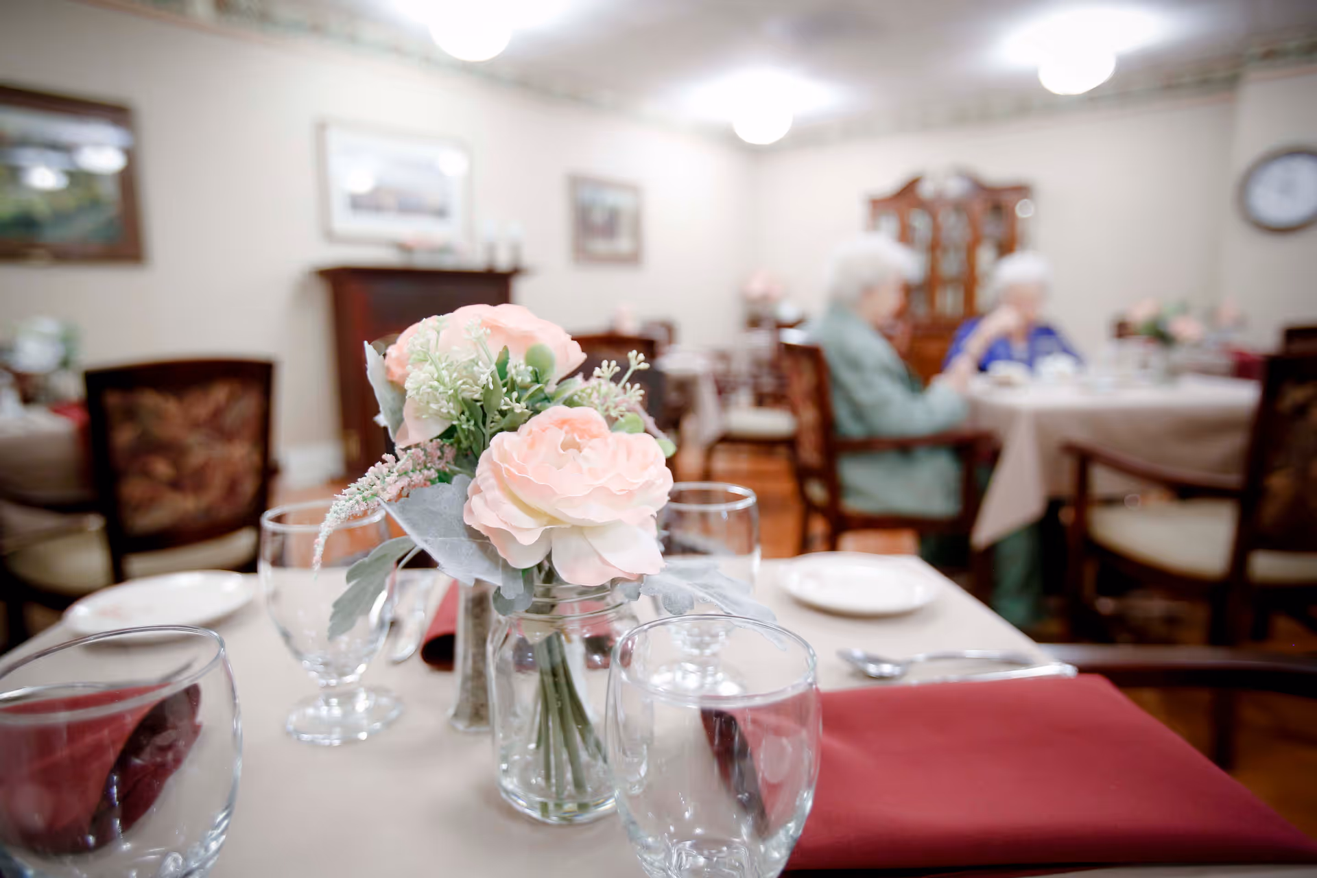 A dining table set with glassware, plates, silverware, and a centerpiece of pink flowers in a glass jar. In the background, two elderly individuals are seated at another table in a softly lit dining room with wooden furniture and framed pictures on the walls.