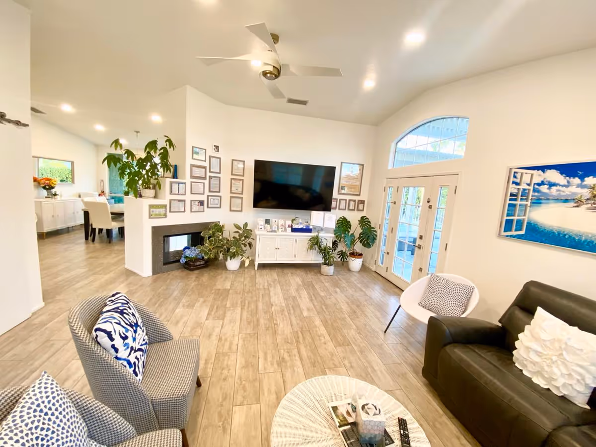 A bright and spacious living room with light wood flooring, a ceiling fan, and a large flat-screen TV mounted on the wall above a white cabinet. The room features several potted plants, a fireplace, and multiple framed pictures on the wall. There are comfortable seating options including a black leather sofa with a white decorative pillow, a white modern chair with a patterned cushion, and two gray armchairs with blue and white patterned pillows. French doors with glass panes lead outside, and a large beach-themed picture hangs on the wall.