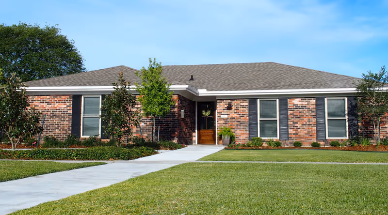Single-story brick building with a gray shingled roof, several windows with black shutters, a front door with a wreath, surrounded by small trees and shrubs, and a concrete walkway leading to the entrance under a clear blue sky.