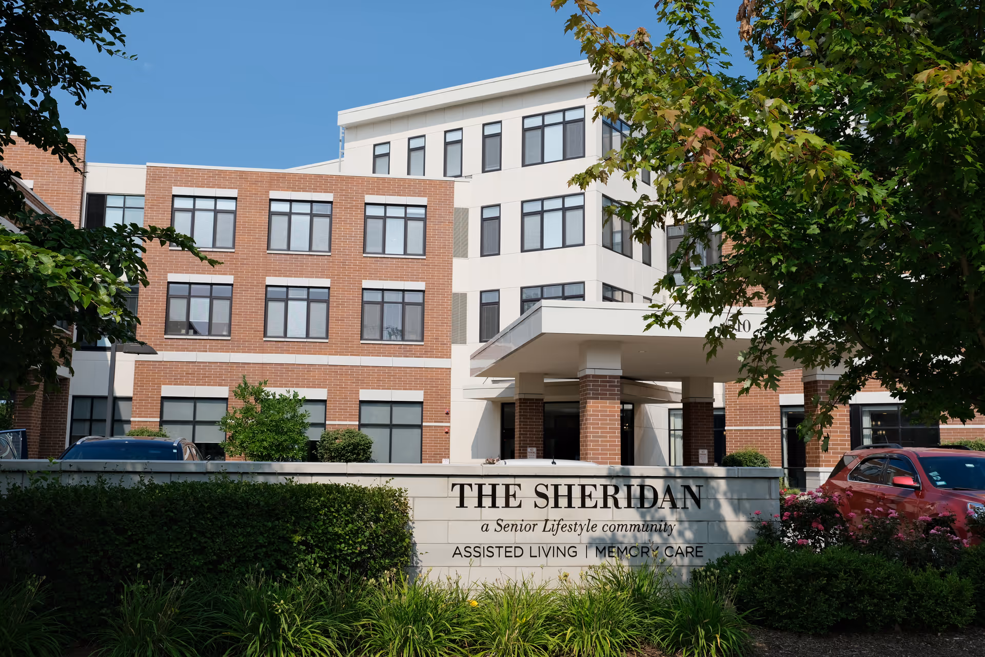 Front entrance of The Sheridan senior living building with a prominent sign, landscaping, and parked cars.