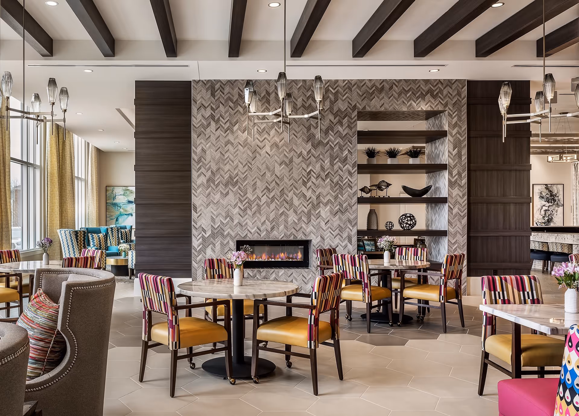 A modern and stylish common area with multiple round tables and colorful chairs featuring striped backs and yellow seats. The room has a decorative fireplace set in a patterned wall, shelves with ornamental items, and large windows with yellow curtains letting in natural light. The ceiling has exposed dark wooden beams and contemporary light fixtures.