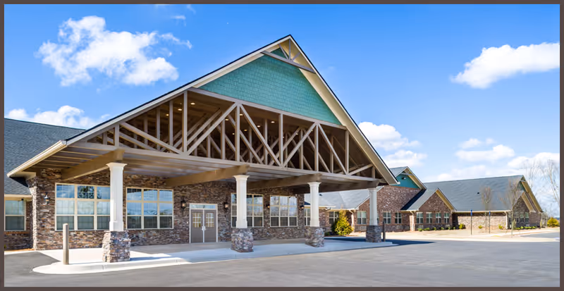 Exterior view of a senior living facility named Antebellum James Burgess featuring a large covered entrance with stone pillars and a peaked roof, set against a blue sky with some clouds.