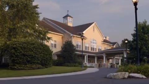 Front exterior of a large beige senior living building with a covered entrance, landscaped grounds, and lampposts.
