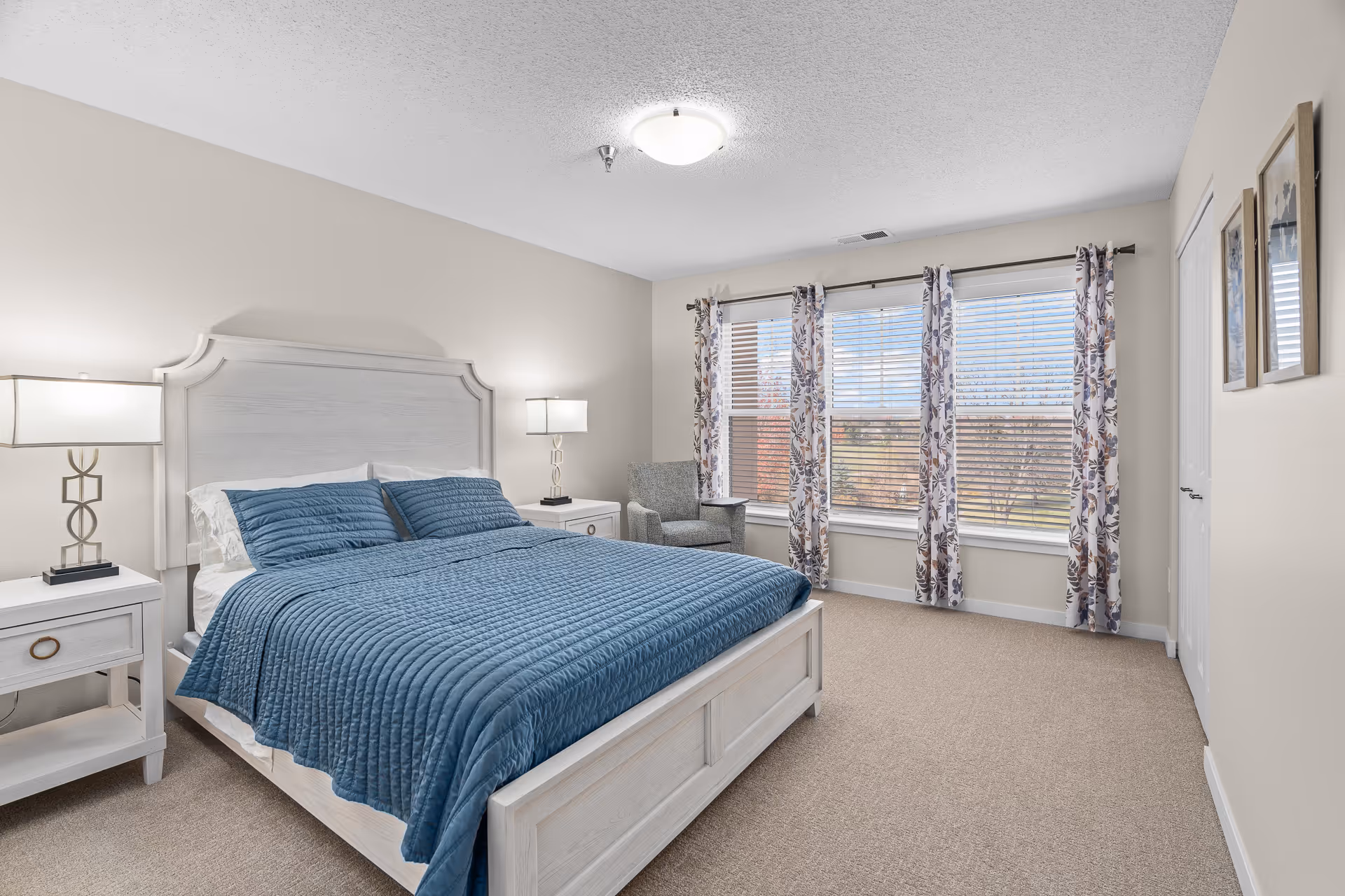 Bright bedroom with a white wooden bed covered in a blue quilt, two nightstands with lamps, an armchair, and large windows with patterned curtains.