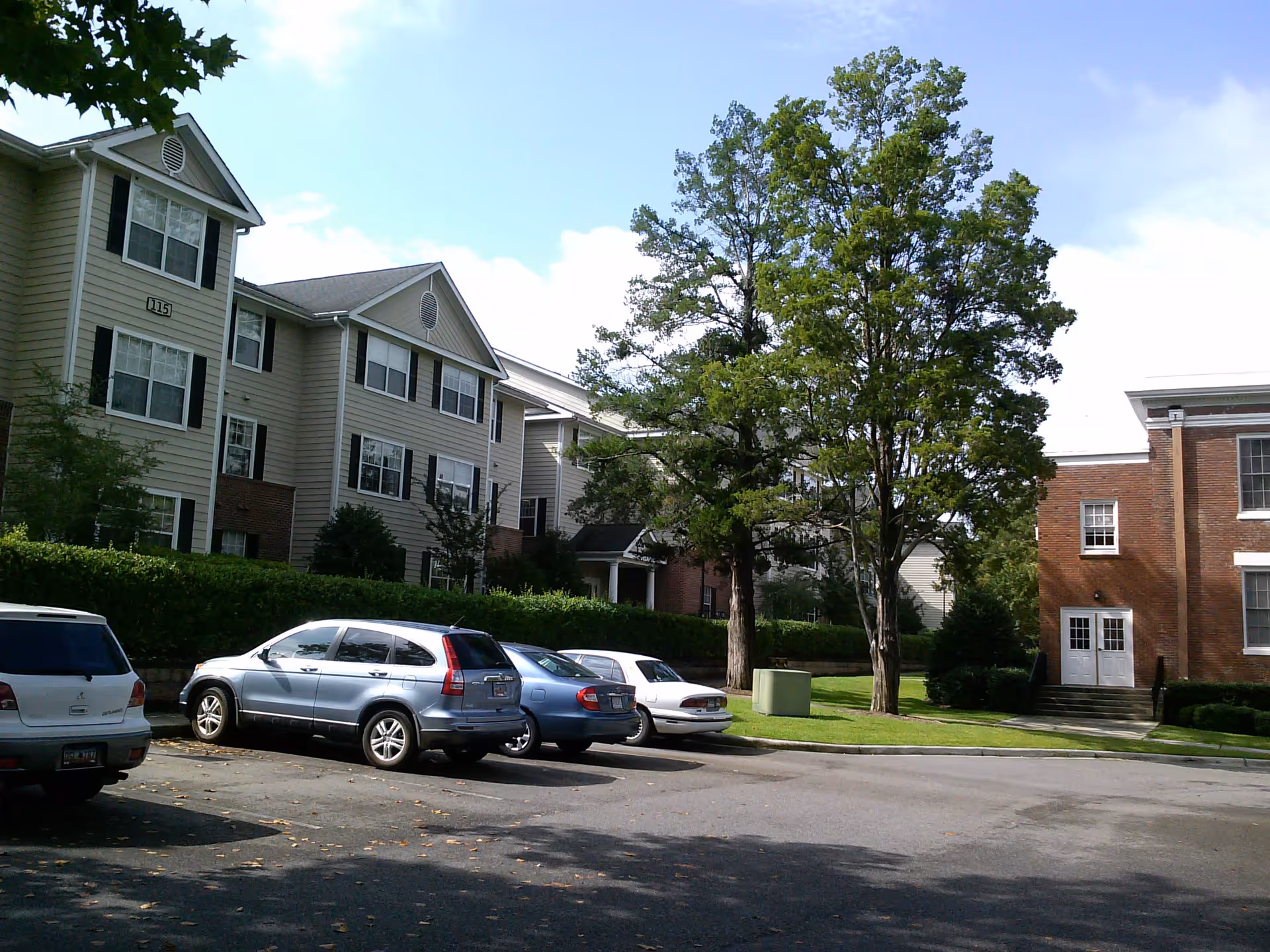 Parking lot with several cars parked in front of a multi-story residential building with beige siding and black shutters, next to a brick building with white trim. There are green trees and a grassy area between the buildings under a partly cloudy sky.