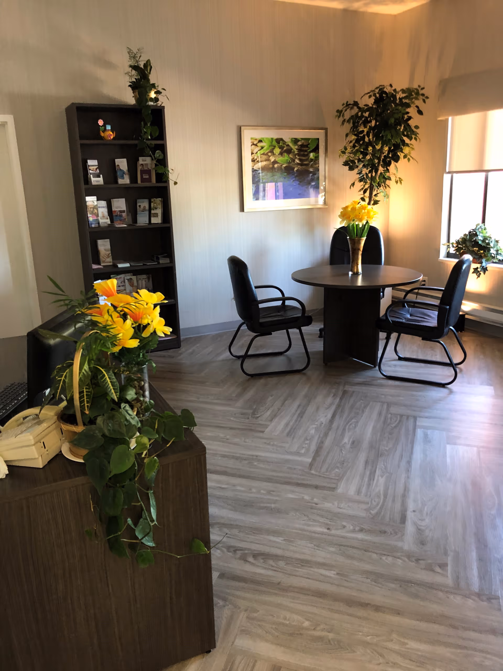 A cozy interior room with a round table surrounded by four black chairs. There are several potted plants, including a tall leafy plant in the corner and a vase with yellow flowers on the table. A dark wooden bookshelf with brochures and decorative items is against the wall, and a framed picture of stacked stones and green leaves hangs above the table. The room has light-colored walls and wood-patterned flooring, with natural light coming through a window with a roller shade.