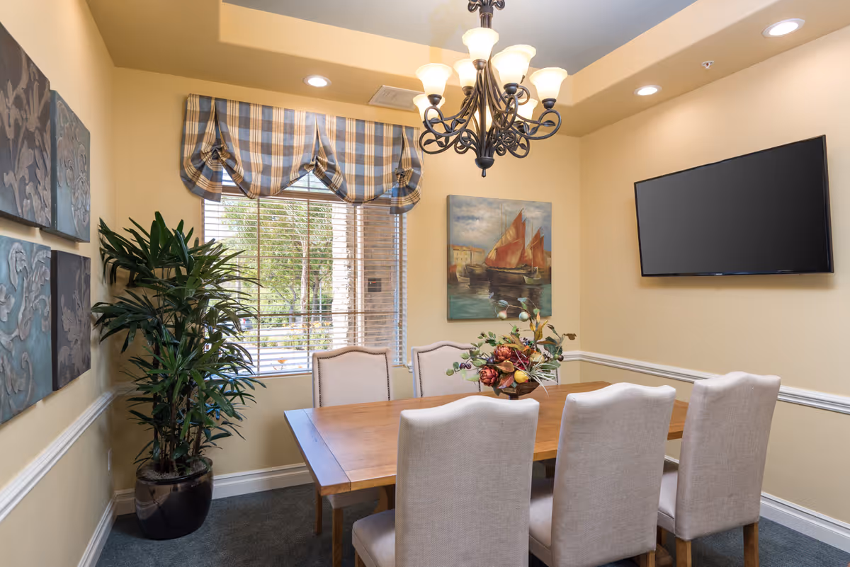 A dining room with a wooden table surrounded by six beige upholstered chairs. A decorative floral centerpiece is on the table. The room has yellow walls with white trim, a window with plaid valance curtains, a potted plant in the corner, a painting of sailboats on the wall, and a flat-screen TV mounted on another wall. A chandelier with multiple lights hangs from the ceiling.