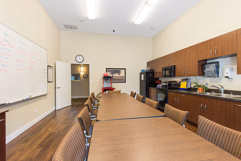 A long meeting or dining room with a wooden table and striped chairs arranged around it. The room features a kitchenette with wooden cabinets, a microwave, a stove, a refrigerator, and a sink. A whiteboard with writing is mounted on the left wall, and a clock is above an open door at the far end of the room. The floor is wooden, and the walls are painted a light beige color.