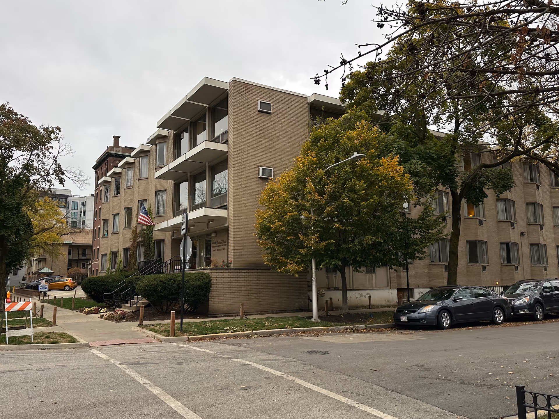 Exterior view of a multi-story brick building with large windows, an American flag near the entrance, and trees with green and yellow leaves surrounding the building. Several cars are parked along the street in front of the building under an overcast sky.