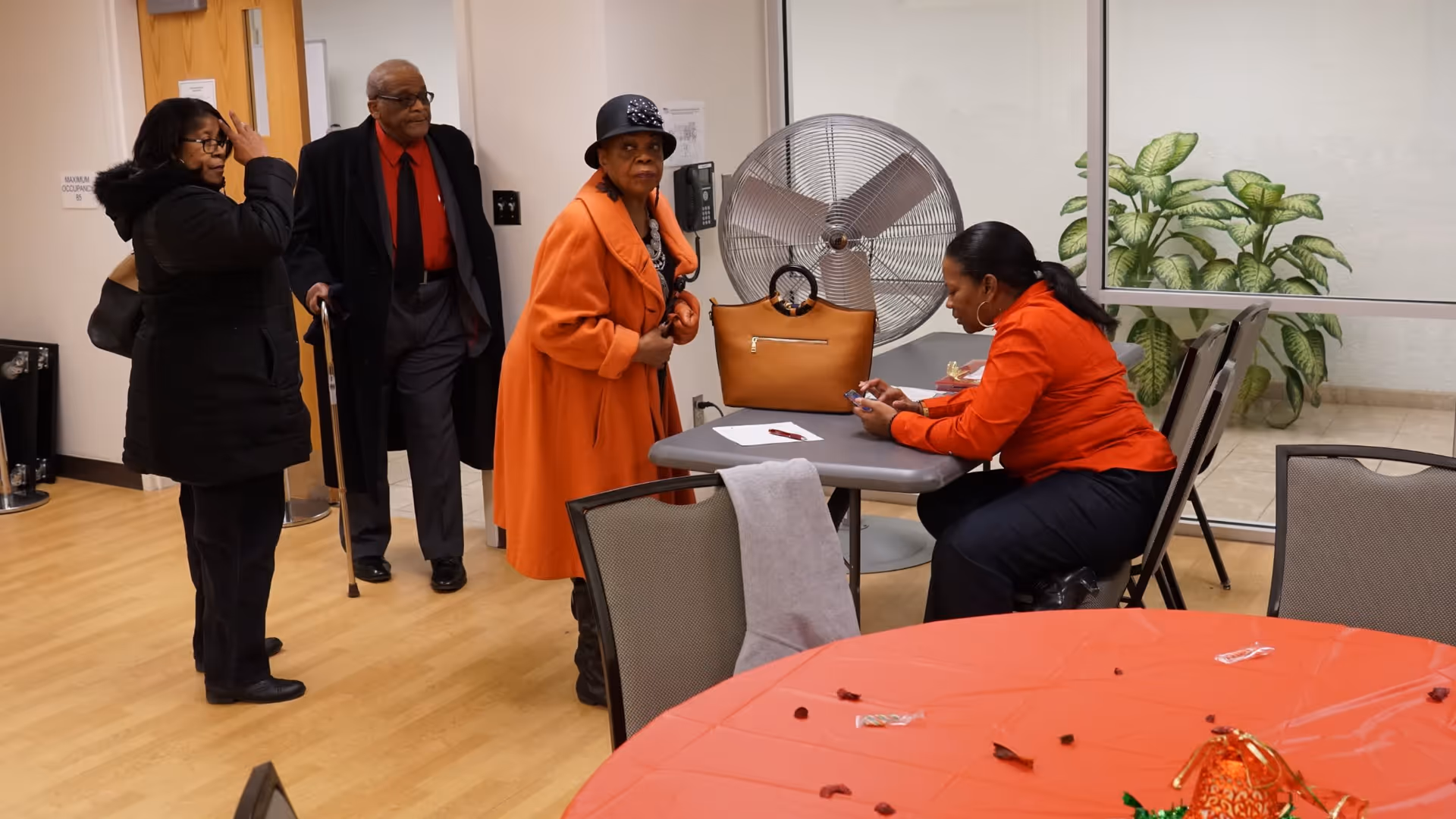 A group of four adults in a room with wooden flooring. Three people are standing near a door, including a man with a cane and two women, one wearing an orange coat and hat. Another woman in an orange shirt is sitting at a table looking at her phone. The room has chairs, a large fan, and a potted plant near a glass partition.