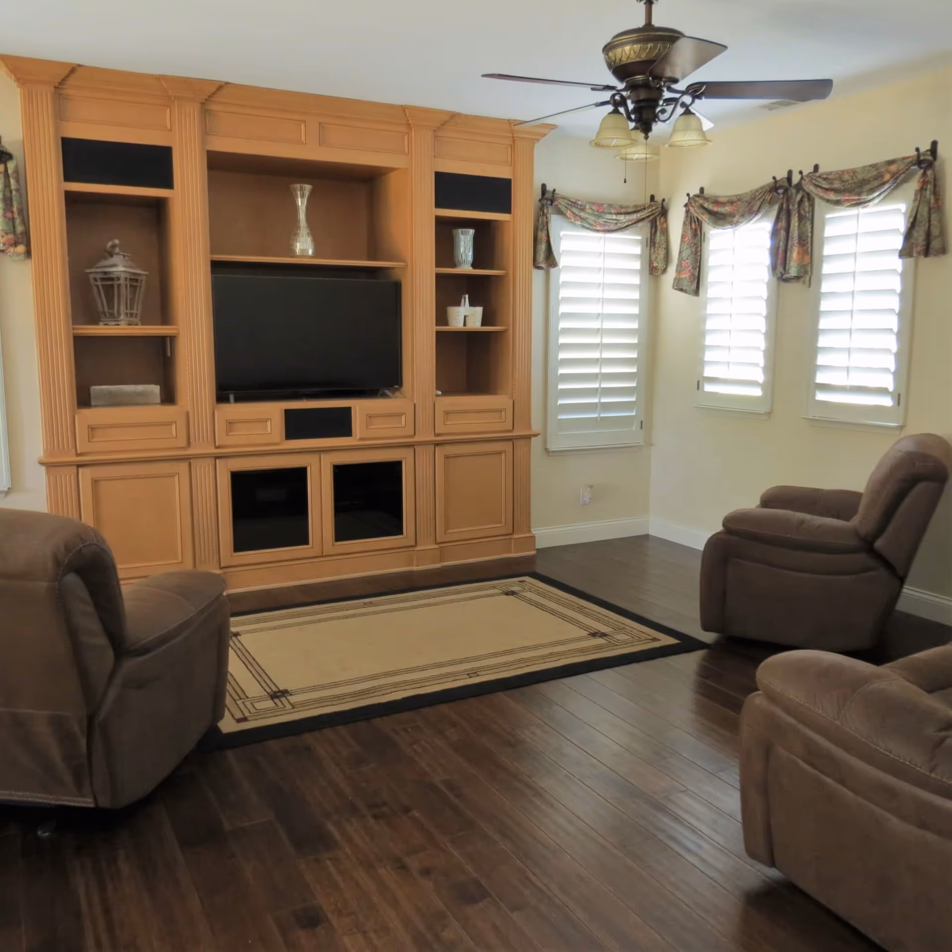 A cozy living room with three brown recliner chairs arranged around a beige area rug on dark wooden flooring. A large wooden entertainment center with shelves and a flat-screen TV is against the wall. Three windows with white plantation shutters and floral valances let in natural light. A ceiling fan with lights hangs from the ceiling.