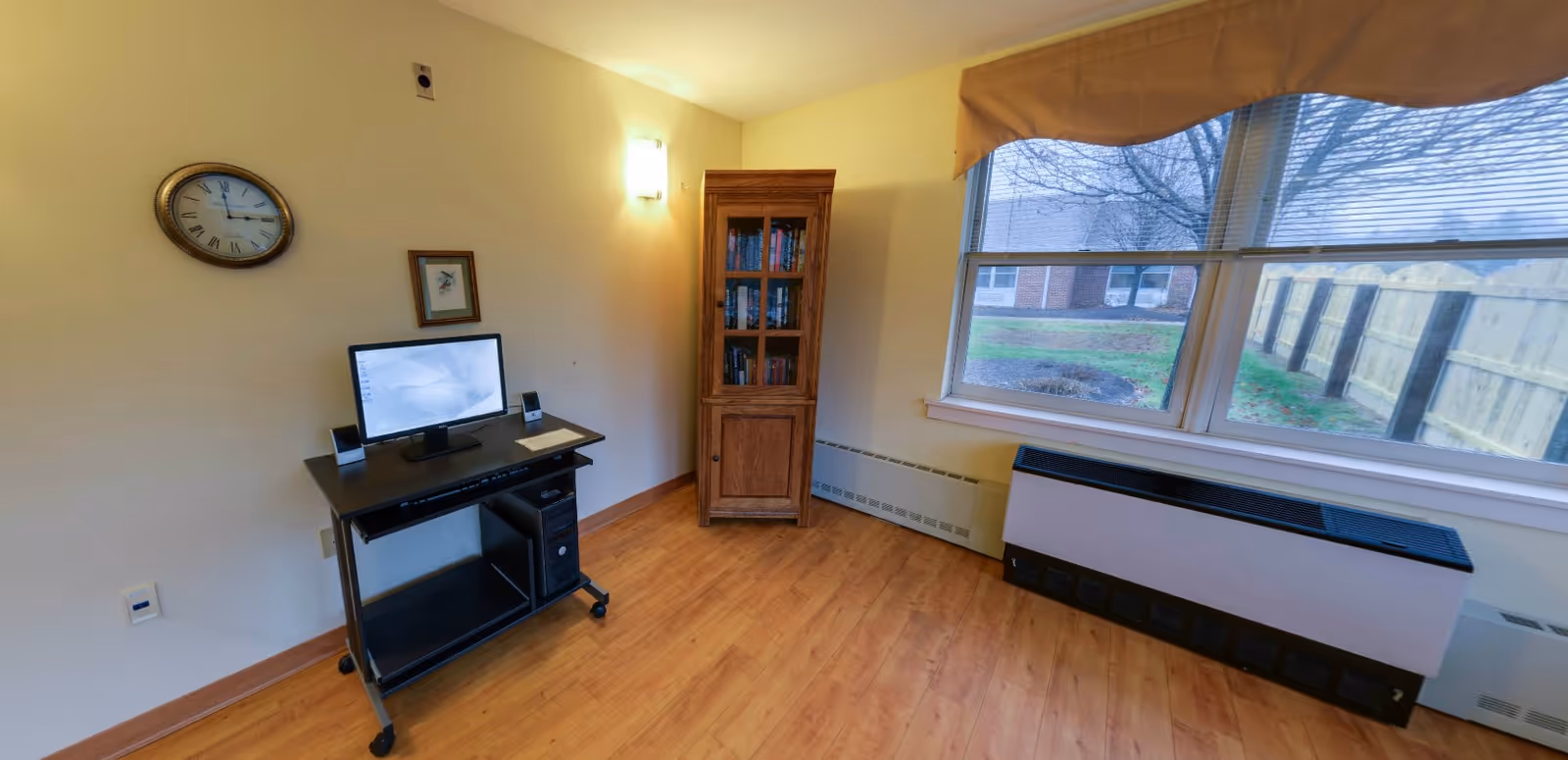Small interior room with a computer desk and monitor, wooden bookshelf, wall clock, and a large window overlooking a fenced yard.