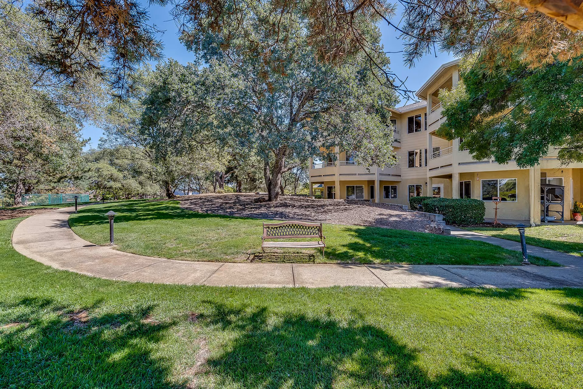 Outdoor view of a senior living facility showing a curved concrete pathway, green grass, a wooden bench, large trees providing shade, and a multi-story beige building with balconies in the background under a clear blue sky.