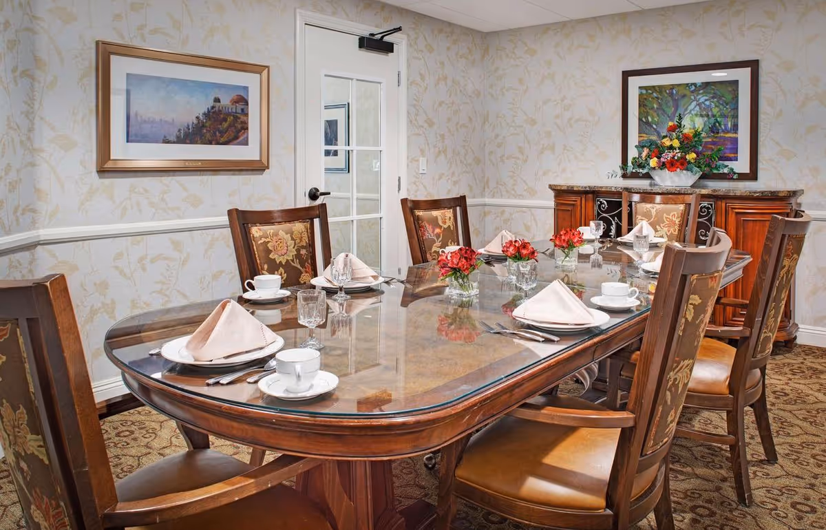 Elegant dining room with a glass-topped wooden table set for eight, napkins and china, floral centerpieces, and upholstered chairs.