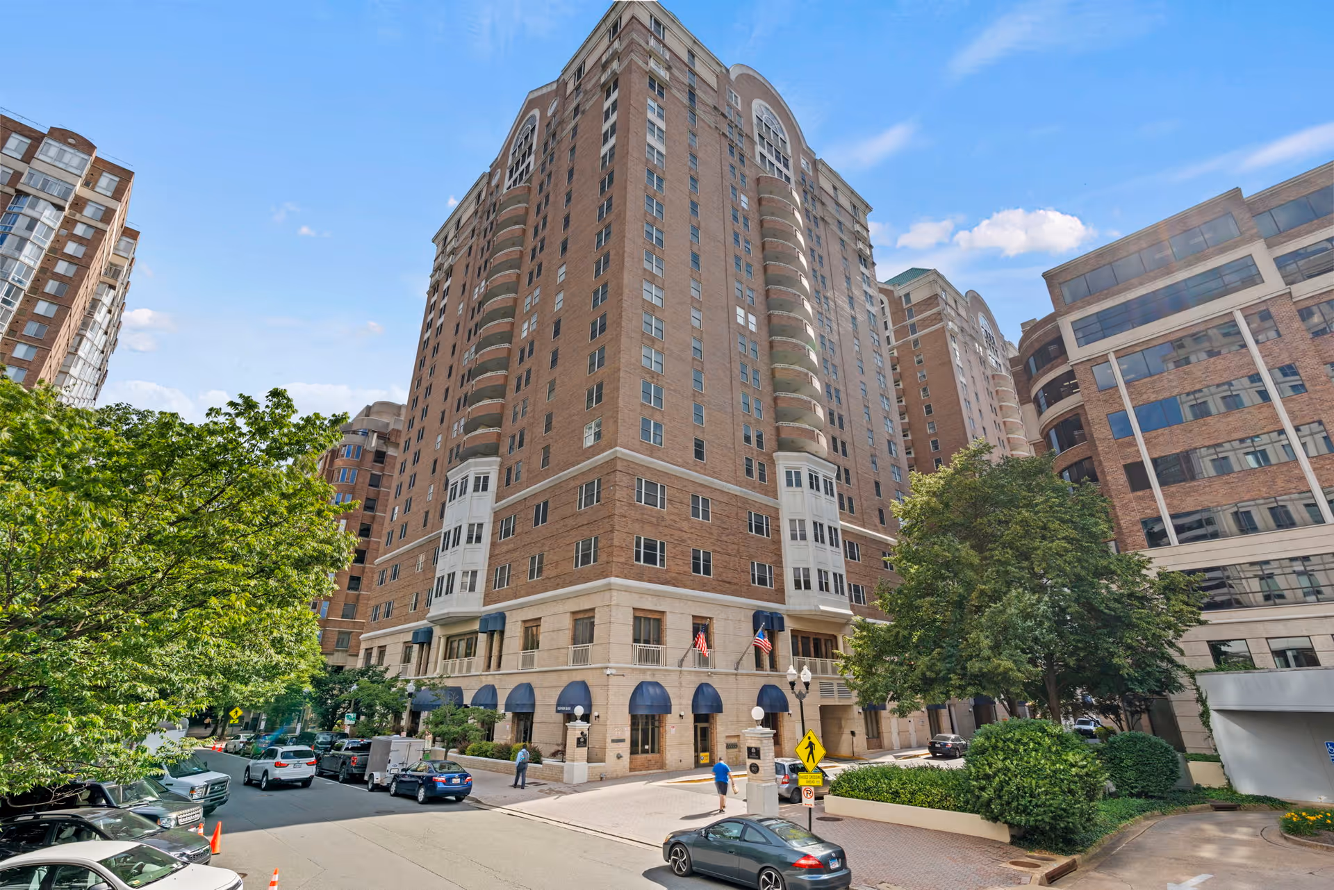 Exterior view of a tall brick residential building with multiple floors and balconies, surrounded by trees and parked cars on the street. The building has blue awnings over the entrance and American flags displayed near the doorway.