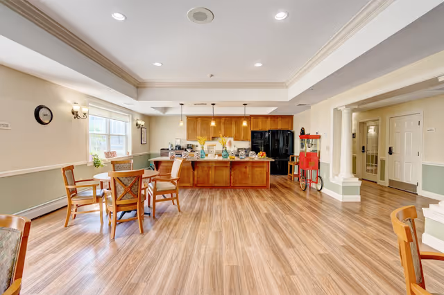 A spacious dining area with wooden floors, several round tables with chairs, and a kitchen area in the background featuring wooden cabinets, a black refrigerator, and a popcorn machine. The room has large windows letting in natural light, recessed ceiling lights, and light green and white walls with decorative molding.