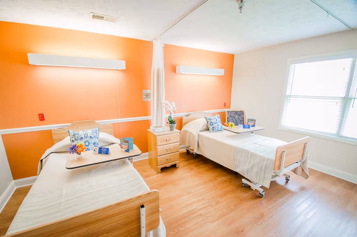 A bright and clean room with two hospital-style beds separated by a small wooden nightstand with drawers. Each bed has a beige blanket, a decorative pillow, and a small overbed table with flowers and cards. The walls are painted orange and white, and there is a large window letting in natural light.