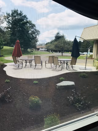 View through a window screen of an outdoor patio with round tables, chairs, umbrellas, and landscaped grounds.