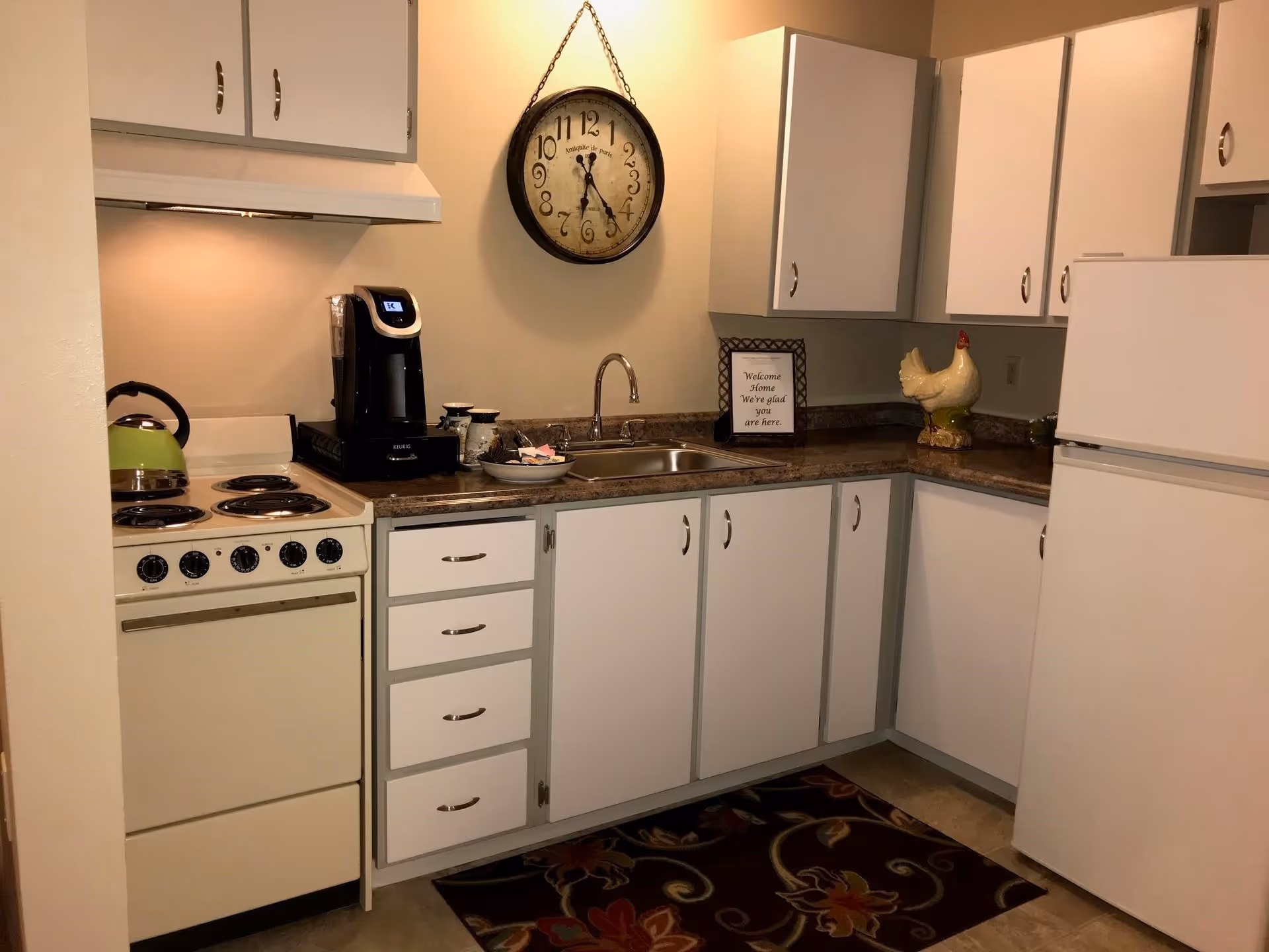 A compact kitchen with white cabinets and drawers, a beige electric stove with four burners, a green kettle, a black coffee maker, a double sink, a wall clock showing 6:25, and a decorative ceramic chicken on the countertop. There is a small framed sign on the counter that reads 'Welcome Home We're glad you are here.' A floral-patterned rug is on the floor in front of the sink.