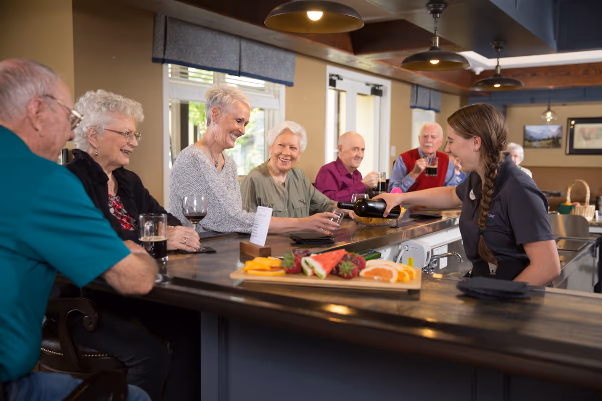 A smiling staff member pours wine for several elderly residents seated at a community bar with a fruit platter on the counter.