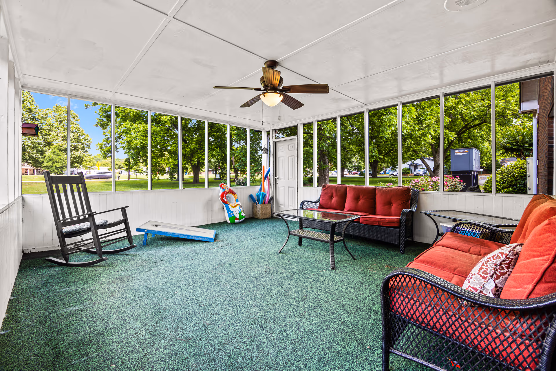 A screened-in porch area with green carpet flooring, a ceiling fan with light, two black wicker sofas with red cushions, a black rocking chair, a glass coffee table, and some pool noodles and a colorful inflatable toy in a basket near a white door. Outside the screened windows, green trees and a grassy area are visible.