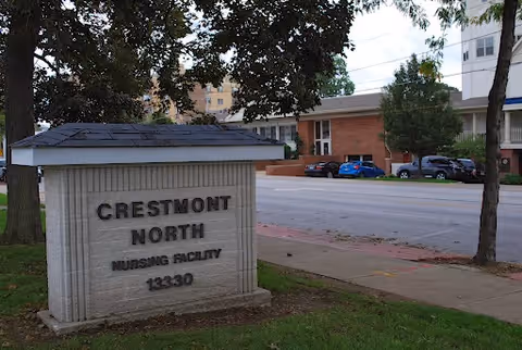 A stone sign reading "Crestmont North Nursing Facility 13330" on a lawn with the facility building and parked cars across the street.