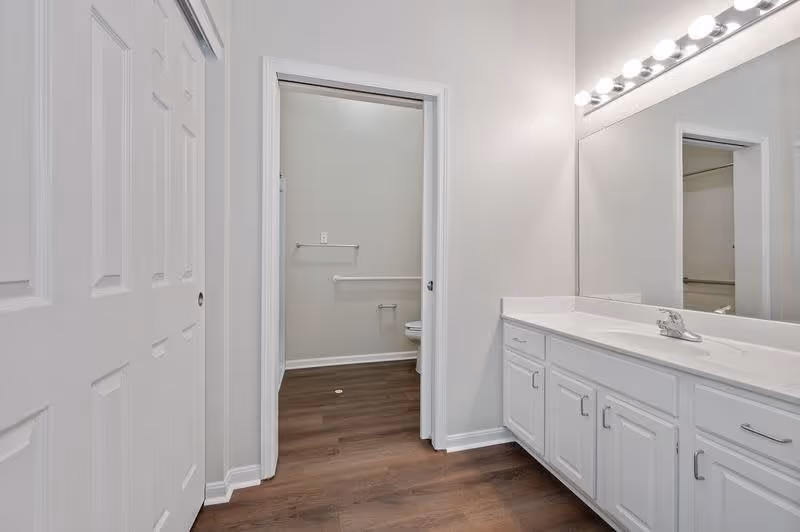 A clean bathroom interior with white walls and cabinetry, a large mirror with lights above a double sink vanity, wood flooring, and a doorway leading to a toilet area with grab bars.