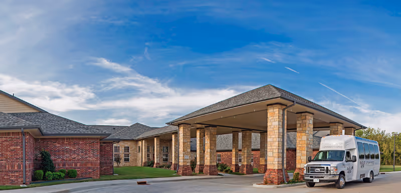 Exterior view of a single-story brick building with a covered entrance supported by stone pillars. A white shuttle bus is parked under the covered entrance. The sky is blue with some clouds.