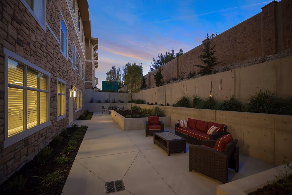 Outdoor patio area at dusk with a stone building on the left, concrete retaining walls on the right, and outdoor seating including a sofa and chairs with red cushions. There are plants along the walls and a small table with chairs in the background.