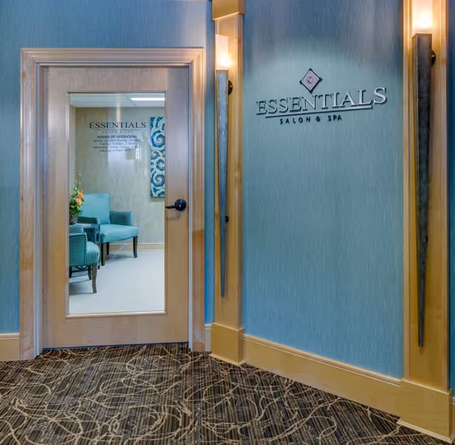 Entrance to Essentials Salon & Spa with a wooden door featuring a glass panel. Inside, two teal upholstered chairs are visible against a patterned wallpaper. The wall next to the door has the Essentials Salon & Spa sign mounted on a blue textured surface with two vertical wall sconces providing light.