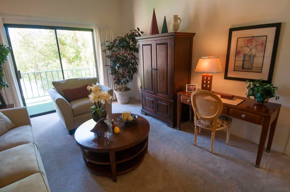 A cozy living room with a beige sofa and armchair, a round wooden coffee table with decorative items, a wooden cabinet, a desk with a chair and lamp, a framed floral artwork on the wall, and a large sliding glass door leading to a balcony with greenery outside.