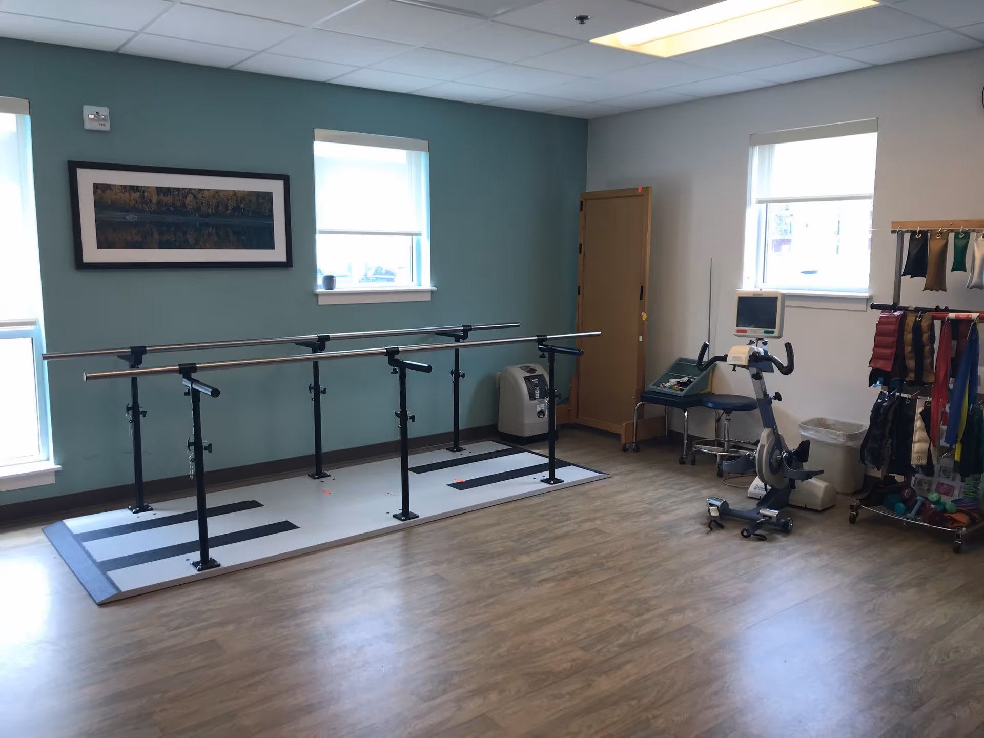 A rehabilitation or physical therapy room with parallel bars for walking exercises, an exercise bike, various therapy equipment, and a wooden floor. The room has two windows with white blinds, a teal accent wall with a framed landscape picture, and light gray walls.