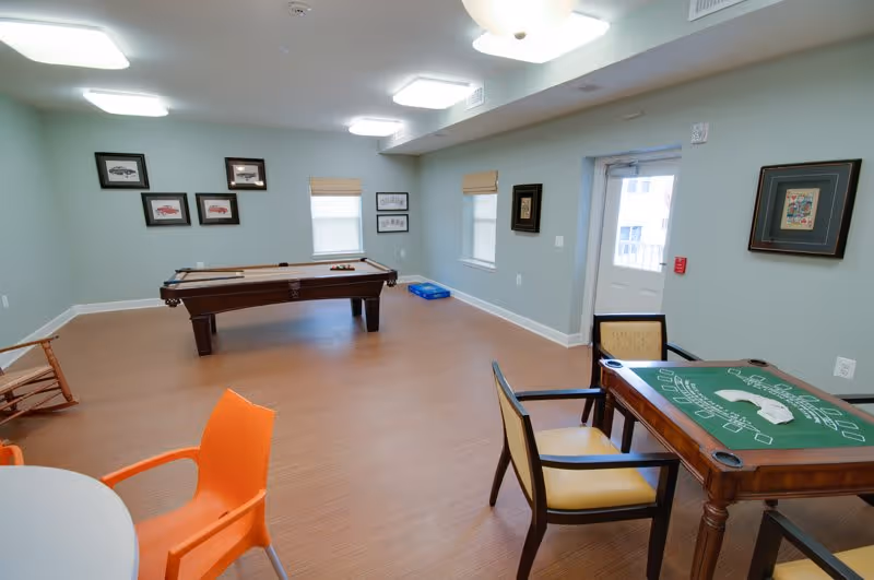 A spacious recreational room with a pool table in the center, a card table with chairs on the right, and an orange chair partially visible on the left. The room has light blue walls with framed pictures, two windows with beige blinds, a door leading outside, and a wooden rocking chair in the corner.